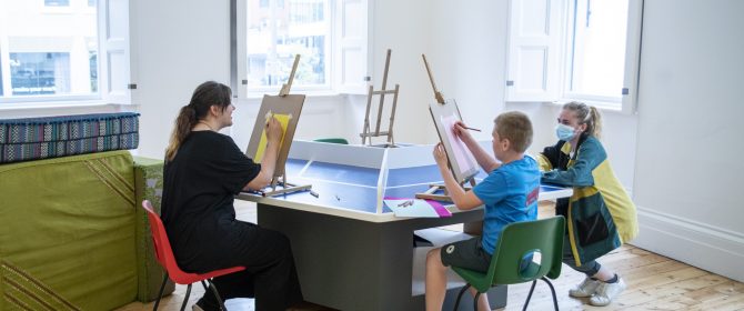 Open Doors Autism friendly workshop - adults and children sat at a table drawing on easels in a bright room with large windows