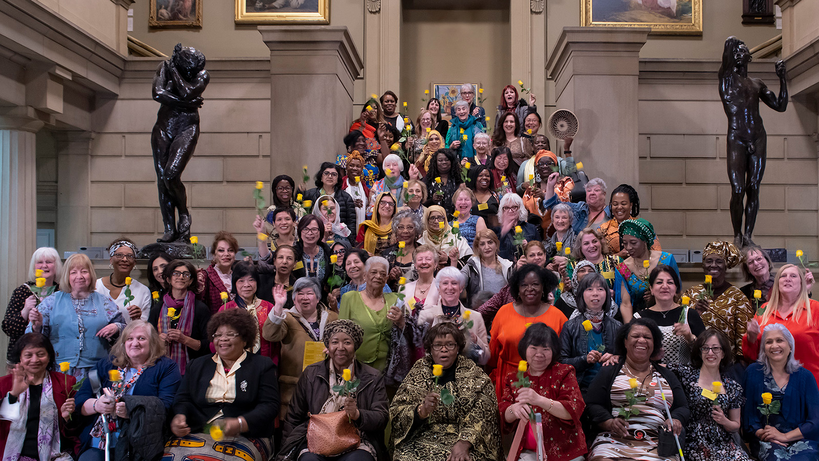 100 women of Uncertain Futures group stood on the stairs in the entrance hall, all holding a single yellow rose