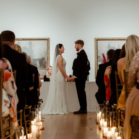 Couple getting married in gallery 16, view in from behind the wedding guests looking towards the ceremony, lights illuminate the isle, bride and groom are holding hands