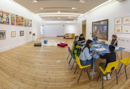 A wide-angle photo of an Open Doors workshop in the Lion's Den. A large white open room with artwork on the walls. Paper and cushions are laid out on the floors and there are adults and children sat at a table taking part in autism friendly activities.