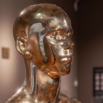 Close up detail of head of a gold male sculpture, the light is shining on his face and you can see the reflection on his cheek