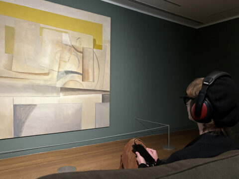 a woman sat down looking at a painting wearing headphones and eye tracking glasses
