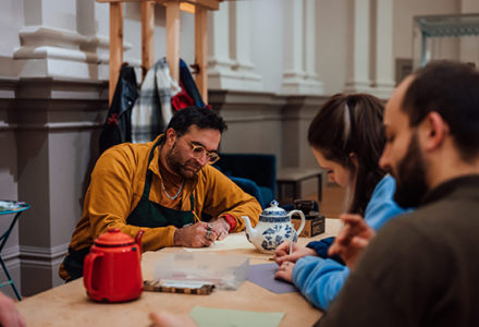 3 people sat around a table drawing objects such as tea pots, in our Trading Station exhibition
