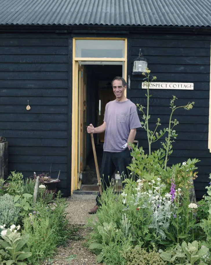 portrait of Derek Jarman holding a rake and stood is his garden in front of Prospect Cottage