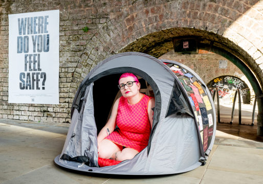 Photo by the artist Gemma inside a camping tent, open and positioned on the sidewalk. On the left a poster that says: Where do you feel safe? The artist wears a fuchsia sleeveless dress that recalls the colour of her hair.