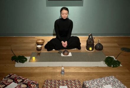 Woman sat crossed legged under a painting in Room to Breathe, a tea ceremony it set up around her with Chinese tea bowls and a tea pot and candles, there are cushions set out around her creating a comfortable atmosphere