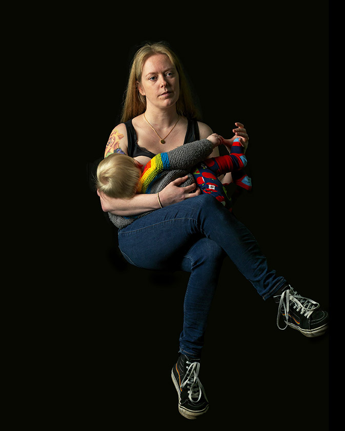 A mother sits with one leg crossed over the other, baby held in her arms breastfeeding. She has long blonde hair, she gazes and something off to the right of the camera. The portrait is set against a background.