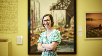 A woman with a short brown bob and glasses wearing a green floral dress, she smiles at the camera with her arms folded in front of a large painting with a yellow background