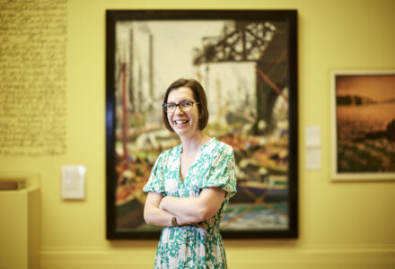 A woman with a short brown bob and glasses wearing a green floral dress, she smiles at the camera with her arms folded in front of a large painting with a yellow background