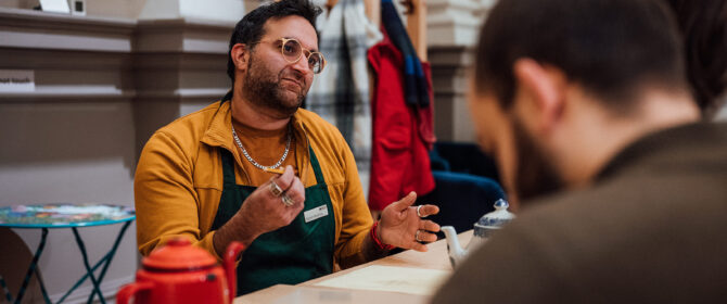 A man in his 30s wearing round glasses and a yellow sweatshirt talking to a couple on the other side of the table, possibly about the teapots sitting in front of them