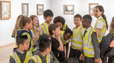 Group of school kids in high vis jackets surrounding a teacher with hands on their eyes. in the background gallery space with artwork of lowry and vallete