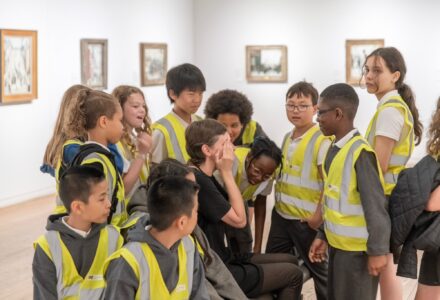 Group of school kids in high vis jackets surrounding a teacher with hands on their eyes. in the background gallery space with artwork of lowry and vallete
