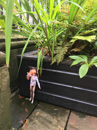 a barbie doll with orange hair is leaning against the planter. on the other scattered foliage