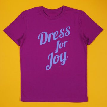 A magenta unisex t-shirt on a yellow background with the slogan Dress for Joy