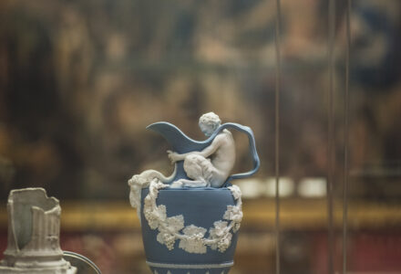 detail of a blue ceramic vase with at the top a satire holding the aperture. In the background an blurred painting.