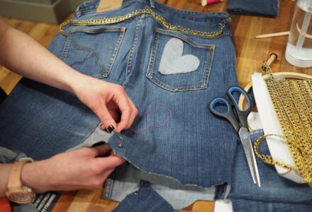 A person customising a denim jacket by sewing a heart patch on the back pocket. Nearby are scissors, gold trim, and sewing materials, indicating a DIY fashion project.